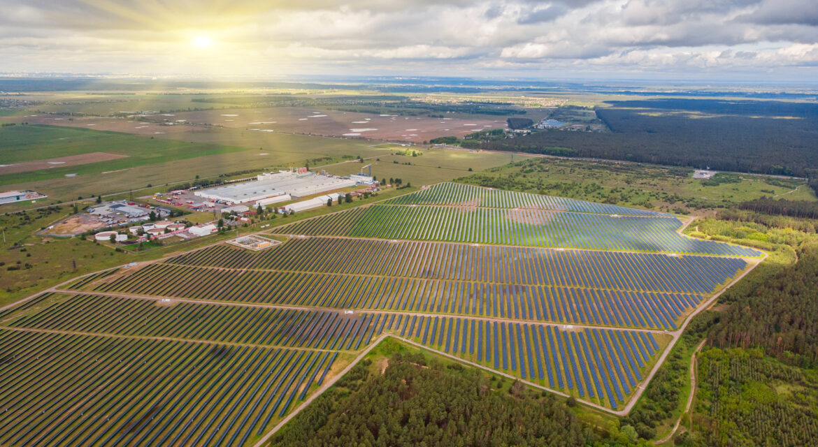 An aerial view of a solar power plant in a vast open field, with the sun shining on a cloudy sky.