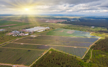 An aerial view of a solar power plant in a vast open field, with the sun shining on a cloudy sky.